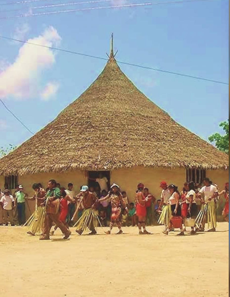 Comunidad indígena en danza ceremonial frente a una gran churuata tradicional de techo cónico bajo cielo despejado